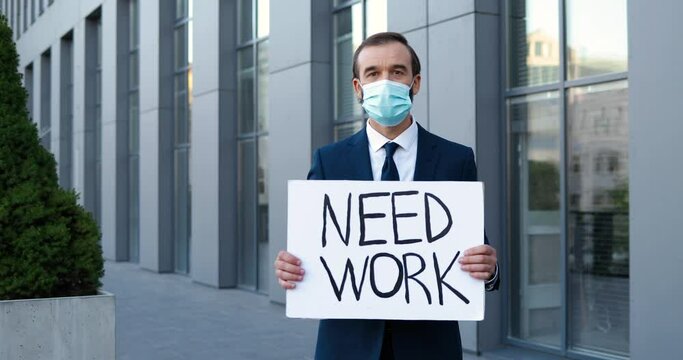 Portrait of retired Caucasian man in medical mask standing outdoor and holding poster Need Work. Senior fired male white-collar worker looking for job during pandemic crisis. Retirement.