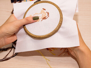 Women's hands hold a white canvas of cross-stitch embroidery in a hoop with a needle, against the background there are multicolored floss threads