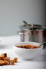 white bowl of thick soup of vegetables and meat, croutons and saucepan on white wooden background vertical image