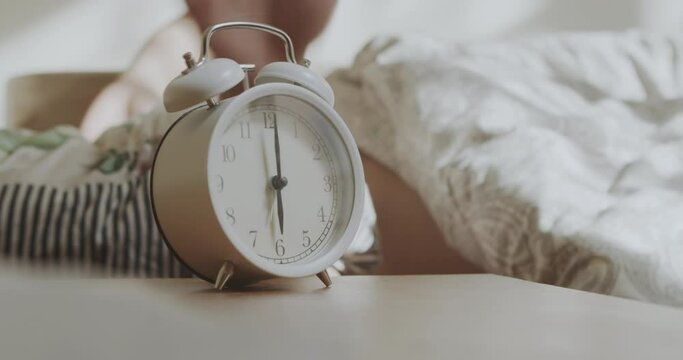 Sleeping man reluctantly wakes up to an alarm clock early in the morning. The man's hand stops the alarm clock to sleep longer.