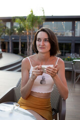 A girl in a yellow skirt and white top sits at a table on the summer terrace of a cafe and hold the cup of coffee. On vacation in Turkey hotel. Smile. Sunny day. Morning. On the sea.
