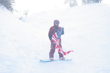 Fototapeta na wymiar winter, leisure, sport and people concept - snowboarder sits high in the mountains on the edge of the slope and looks into the distance. snowboarder holding american flag