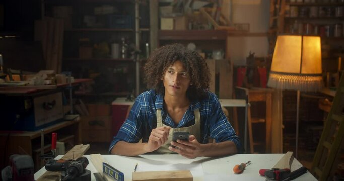 Pleasant curly young carpenter man using mobile device online get inspiration in Pintrest, attractive Latin joiner master working sitting next to work-desk with smartphone connected workshop