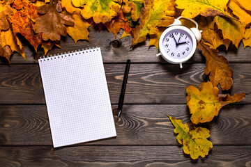 Notepad with a pen among autumn leaves and alarm clock on a brown wooden table