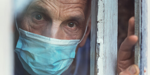 lonely senior man wearing medical face mask looking through window of nursing house