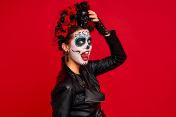 Closeup photo of funny folklore witch, creepy lady calavera with makeup masquerade. Sugar skull girl showing tong, wear floral headwear and leather jacket isolated on red background.