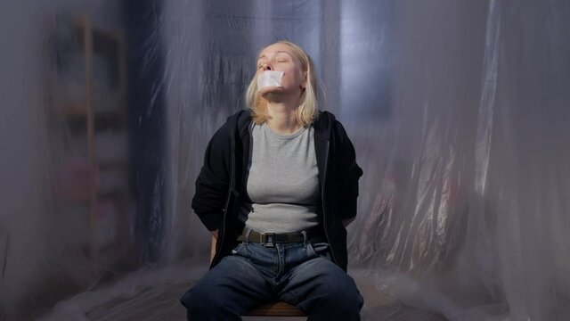 Female hostage sitting on a chair in a room with her mouth taped shut