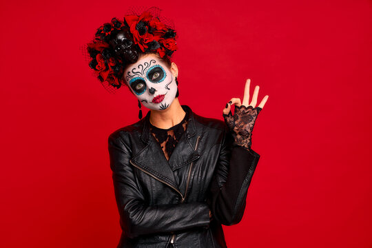 Romantic dead gir with sugar skull makeup with a wreath of flowers on her head and skull, wearth black gloves showing OK gesture isolated on red background. concept of Halloween or La Calavera Catrina