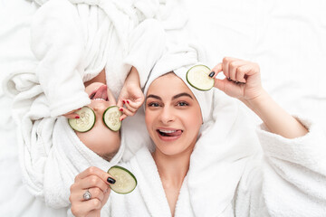 Positive mother and daughter doing face mask applying pieces of cucumber to their eyes, wrapped in a white bath towel, beauty procedures at home. Facial skin care, cosmetology and spa