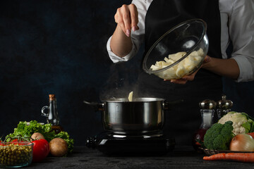 The professional chef pours raw potato from glass bowl into the pot with boiling water on dark blue background. Backstage of preparing meal at the restaurant kitchen. Traditional dish for dinner.