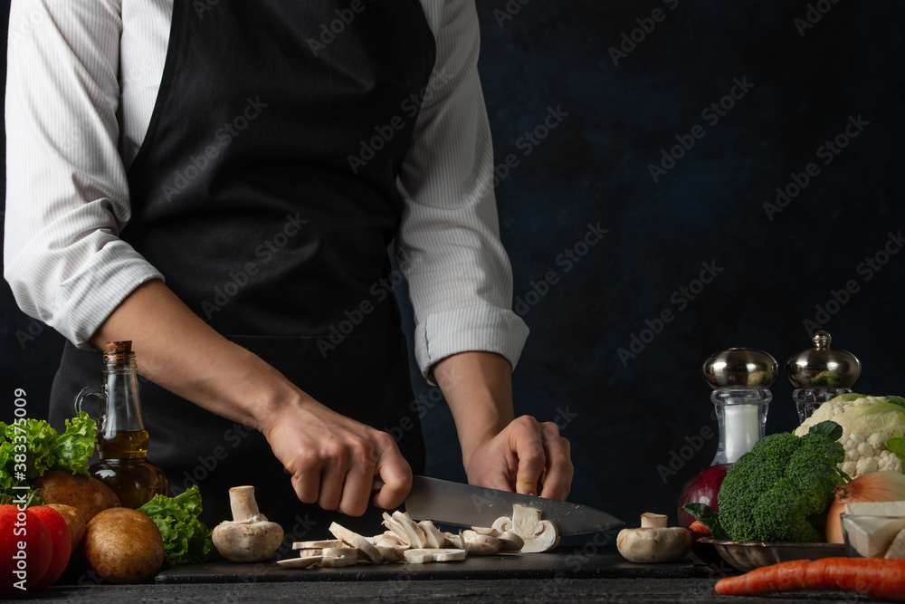 Wall mural the professional chef in black apron cuts with knife mushrooms on black chopped board on dark blue b - Wall murals
