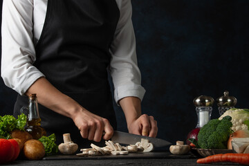 The professional chef in black apron cuts with knife mushrooms on black chopped board on dark blue background. Backstage of preparing restaurant meal for dinner. Food concept. Close-up.