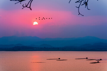 Beautiful nature landscape red sun on colorful sky and birds flock flying in a row over mountain and lake water during sunset, fish farming in cages background at Krasiao Dam, Suphan Buri in Thailand
