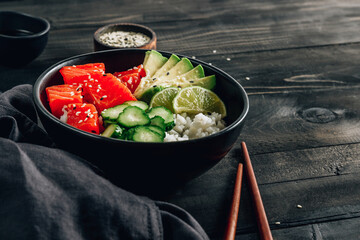 Hawaiian salmon poke bowl with cucumber, avocado and sesame seeds on black wooden table.
