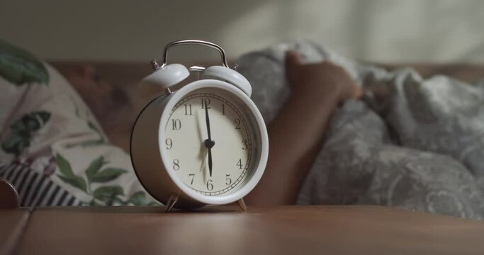 The man is woken up by an alarm clock at six in the morning. The man's hand stops the alarm clock to sleep longer. Sleeping man reluctantly wakes up to an alarm clock early in the morning.