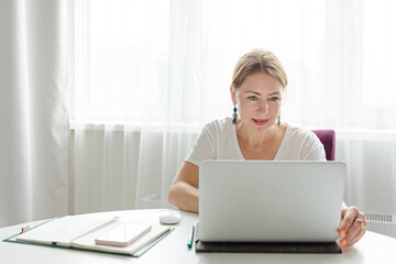 middle-aged woman works on a laptop at home in a light interior at a remote job