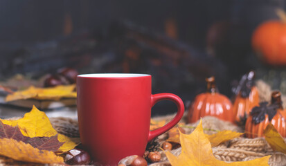 Naklejka na ściany i meble Red mug with tea on the background of a soft, knitted scarf, autumn leaves, pumpkin. Autumn background, cozy evening, fireplace. 