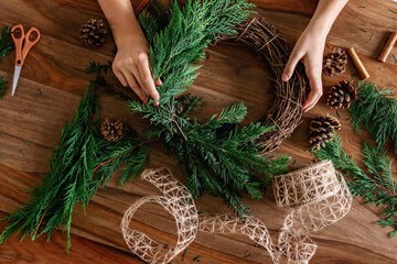 Overhead shot of hands making handmade Christmas wreath with twigs, pine cones, cinnamon sticks,...