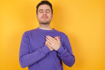 Handsome man with sweatshirt over isolated yellow background smiling with hands on chest with closed eyes and grateful gesture on face. Health concept.
