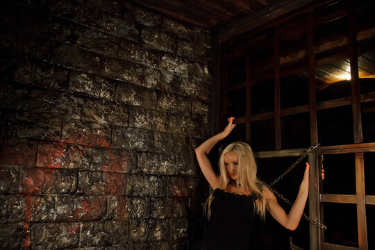 Pretty young sexy blonde woman in black attire in interior of medieval dark room with cages and chains on background of an old texture wall. Image of Queen of Halloween horror night. Copy space