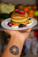 Plato con panqueques listos para comer acompañados de frutas frescas de estación, siendo estas fresa y plátano picados y arándanos, sujetados por una mano con un tatuaje de un obturador