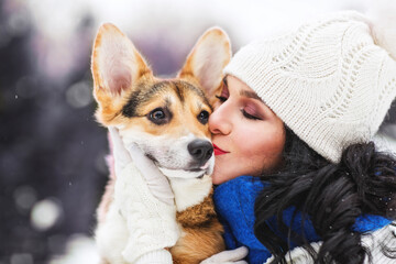 Outdoors lifestyle image of stunning woman hugging and kissing puppy corgi in the park. Beautiful face. Wearing stylish outerwear, white knitted hat and blue scarf. Playing with the dog. Close-up. 