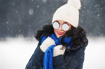 Fototapeta na wymiar Outdoors lifestyle close up portrait of beautiful girl walking in the snowy winter park. Smiling and enjoying wintertime. Wearing stylish mirrored sunglasses, down jacket, knitted hat. It's snowing