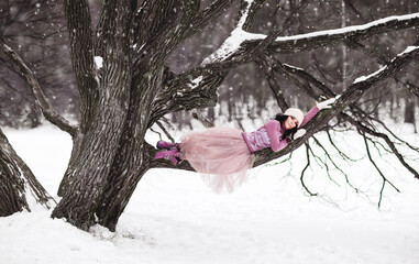 Girl lying on a tree branch. Winter's tale lifestyle portrait of charming young woman enjoying snowfall in snowy park. Wearing long tulle skirt and stylish white knitted hat and gloves. It's snowing. 