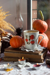 Autumn drink on the windowsill. Lemon tea in a white patterned cup, old books, orange pumpkins, burning candles and a kerosene puddle in the background. Cozy autumn concept