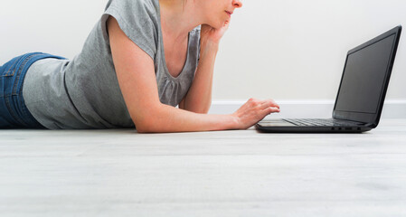 Fototapeta premium Croped caucasian woman lying on floor and working with a laptop. Female in casual clothes with t-shirt and jeans relax at home with internet. Copy space. Banner