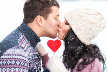 Outdoors lifestyle close up portrait of happy kissing couple in love. Holding in their hands red lollipop in shape of heart. On honeymoon. Wearing knitted hat, mittens and sweaters.  Love concept