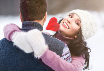 Outdoors romantic portrait of happy hugging couple in love. Girl smiling and holding in her hand red lollipop in shape of heart. Wearing knitted hat, mittens and sweaters. Love concept. Valentine day
