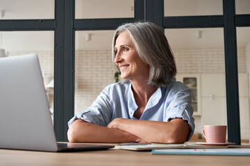 Smiling stylish mature middle aged woman sitting at home office workplace looking away. Happy older...
