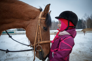 Close up of a little girl jockey smiling and looking to the horse eyes. Girl horse rider with a horse in a winter open riding hall, looking eye to eye. Girl and horse friendship, equestrian sport