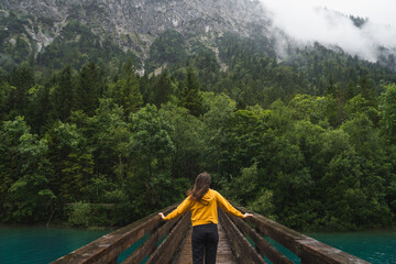 Girl in yellow walking on a bridge over a clear blue river surrounded by green forest