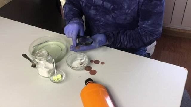 A man in a mask and rubber gloves examines a copper coin and puts it in phosphoric acid. Beside soda and sulfuric ointment for cleaning copper coins.