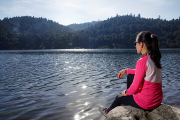 Mujer sentada a la orilla de un lago observando el paisaje del bosque al fondo