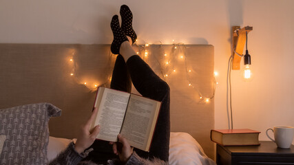 Pasando una tarde de otoño leyendo y descansando. Mujer adulta leyendo un viejo libro cómodamente acostada en una cálida y confortable habitación iluminada tenuemente - obrazy, fototapety, plakaty