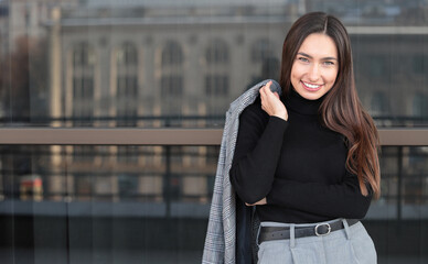 Beautiful smiling woman stand on the urban office building background holds a jacket on her shoulder looking at the camera.Business success concept.Empty space for text or message