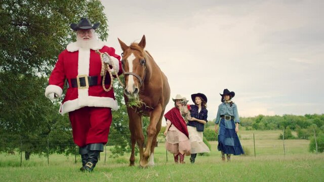 A cowboy Santa Clause walking along a ranch in Texas with a horse by his side and kids following behind him. 