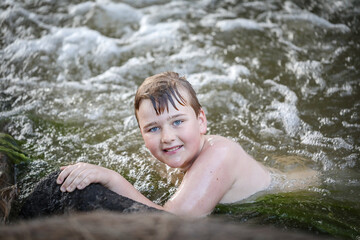 Boy swimming in river at dusk holding onto edge looking up at camera. Moody image with rich tones