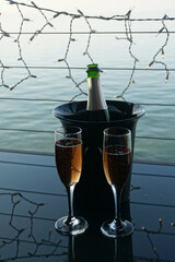 View of an open bottle of pink champagne or sparking wine in a bucket of ice, two glasses, with railing, lights and water in the background