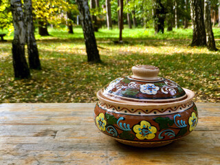 Traditional slavic dishes on the old wooden table, autumn birch forest, ceramic pot with floral...