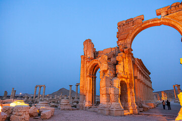 landscape image of the ancient city of Palmyra at night right before sunrise. Ancient columns and arched doors are illuminated with spot lights with night sky in background. Few tourists are walking.