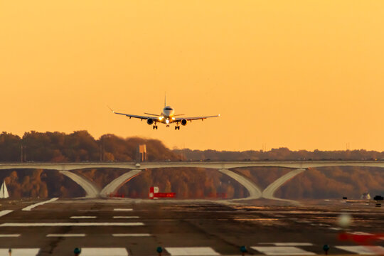 A Passenger Airline Is Slowly Descending To Land On A Runway At Sunset. The Lights Of The Plane Are On And Wheels Are Down. There Is Scenic Sunset Sky, Trees And Bridge In Background.