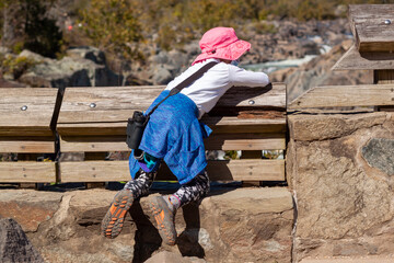 Fototapeta na wymiar A curious little girl wearing pink solar hat and sneakers is climbing on the stone barriers to see the great falls of Virginia. This is a dangerous act with risk of falling. She has binoculars.