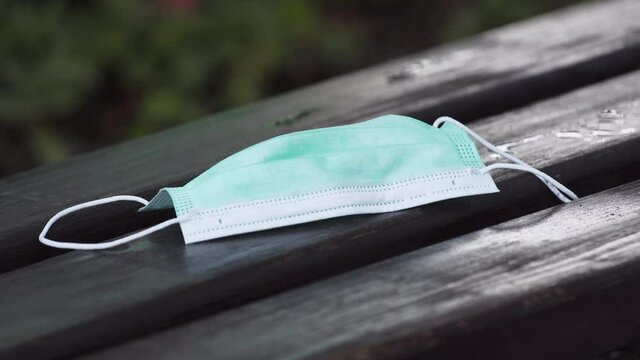 A person putting a simple green protective Covid-19 face mask on a dark wooden bench, picking it up again after a moment, blurry bushes in the background, static close up 4k shot.