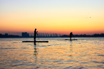 Silhouettes of two men paddle on stand up paddle boarding (SUP) on quiet winter or autumn Danube river at sunrise. Colorful sunset over the river with silhouettes of people