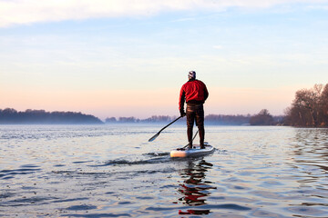 Back view on man rowing on SUP (stand up paddle board) in autumn Danube river at foggy morning...