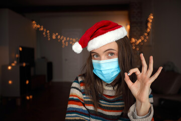 Happy adult female in medical mask and Santa hat gesturing OK during Christmas celebration at home